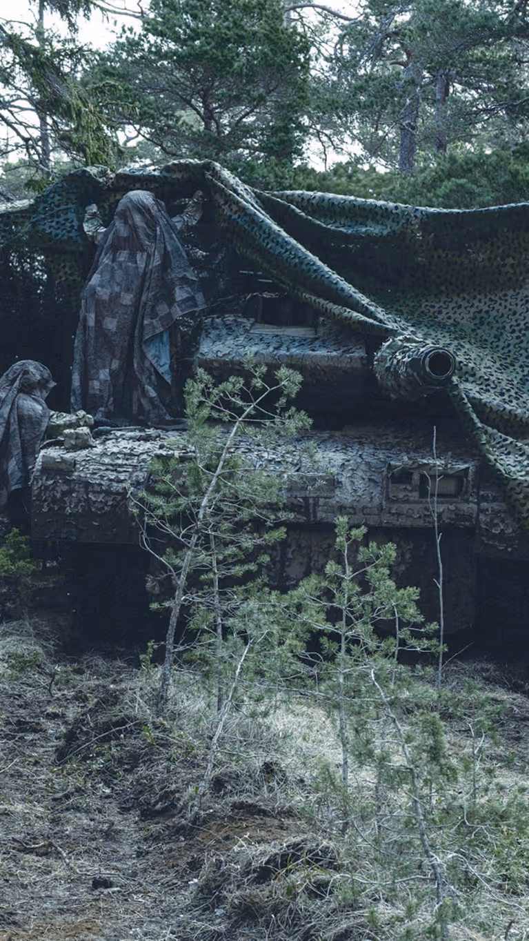 Militair voertuig in bos onder camouflagenet met daaromheen een aantal gecamoufleerde militairen.