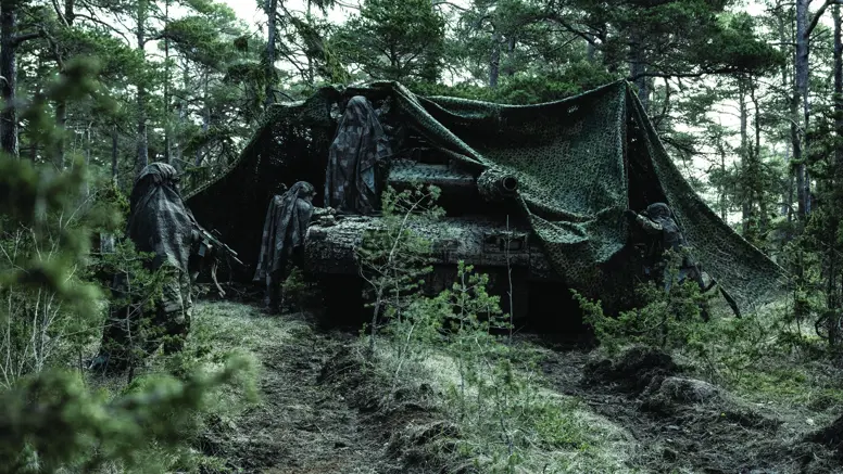 Militair voertuig in bos onder camouflagenet met daaromheen een aantal gecamoufleerde militairen.