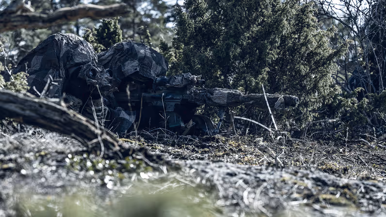 Een gecamoufleerde militair ligt achter zijn wapen in het bos.