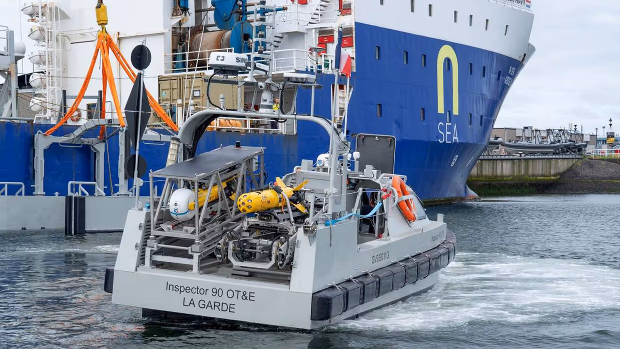 Links een grijs bootje met achterop verschillende drones. Dit vaart langs een groot vrachtschip. Daarnaast een gele, raketvormige drone met voorop een witte bol. Deze is ook te zien achterop het varende bootje.