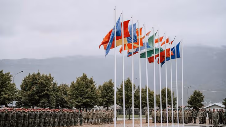 Vlaggen van 11 landen wapperen op lange masten. Daarvoor staan rijen dik militairen.