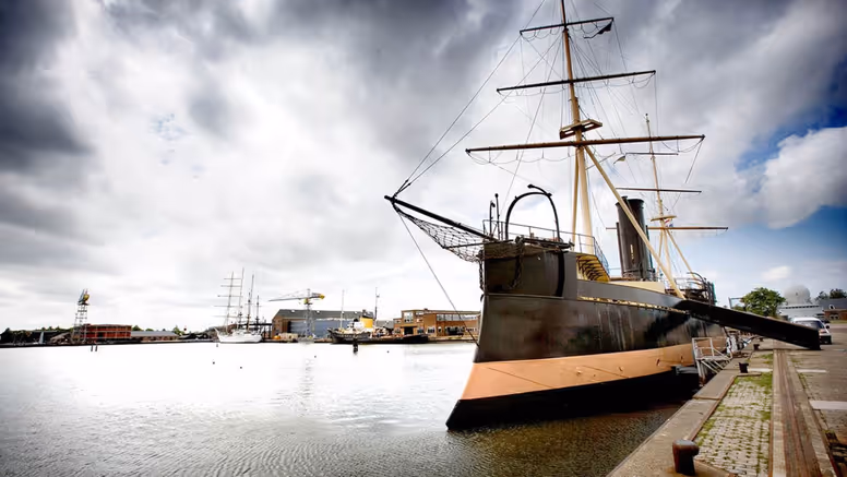 Een groot, donker schip met 2 masten ligt aan de kade in een haven onder een bewolkte lucht.