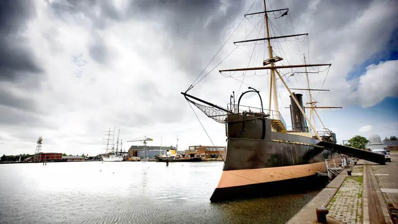 Een groot, donker schip met 2 masten ligt aan de kade in een haven onder een bewolkte lucht.