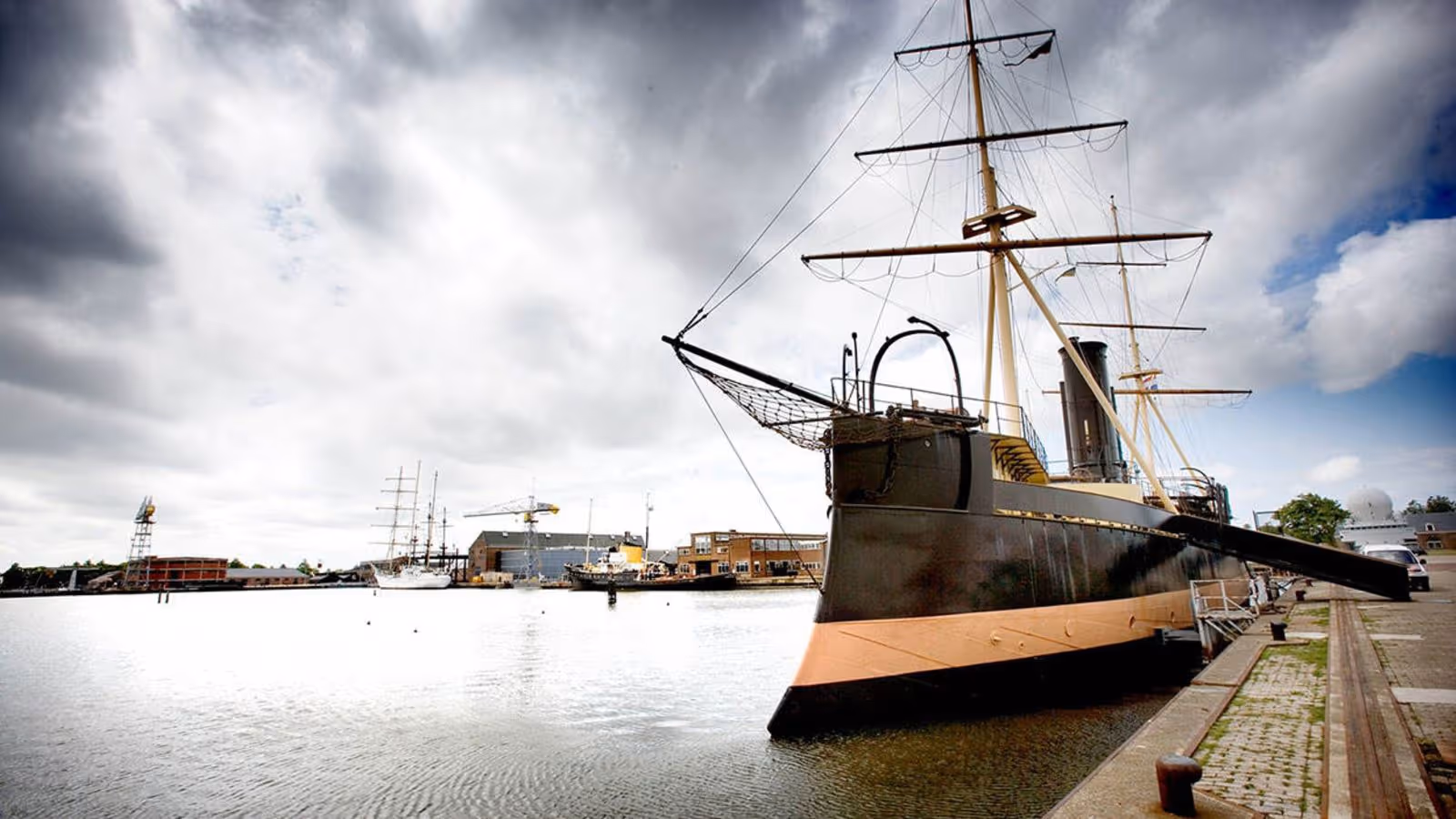 Een groot, donker schip met 2 masten ligt aan de kade in een haven onder een bewolkte lucht.