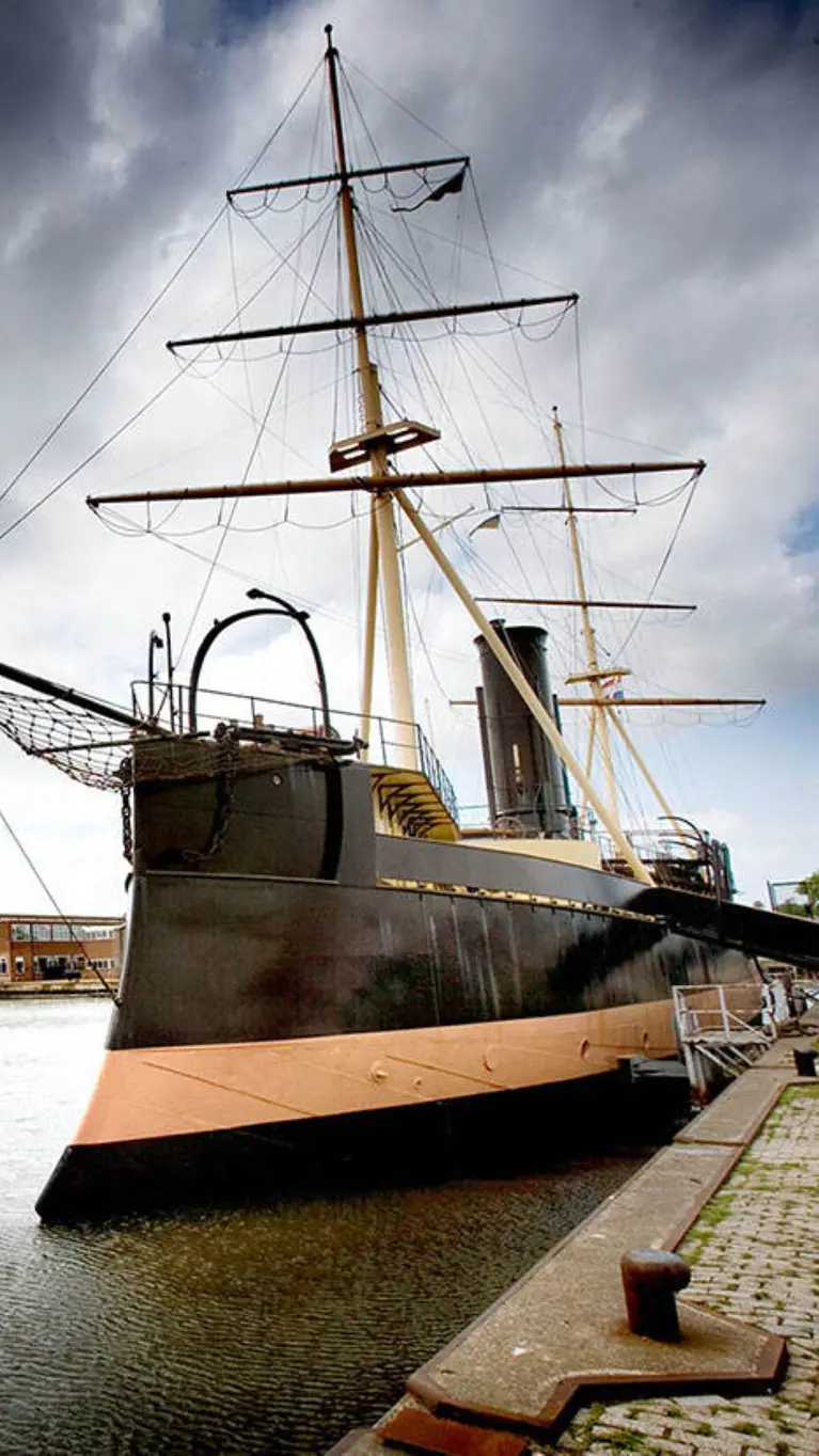 Een groot, donker schip met 2 masten ligt aan de kade in een haven onder een bewolkte lucht.