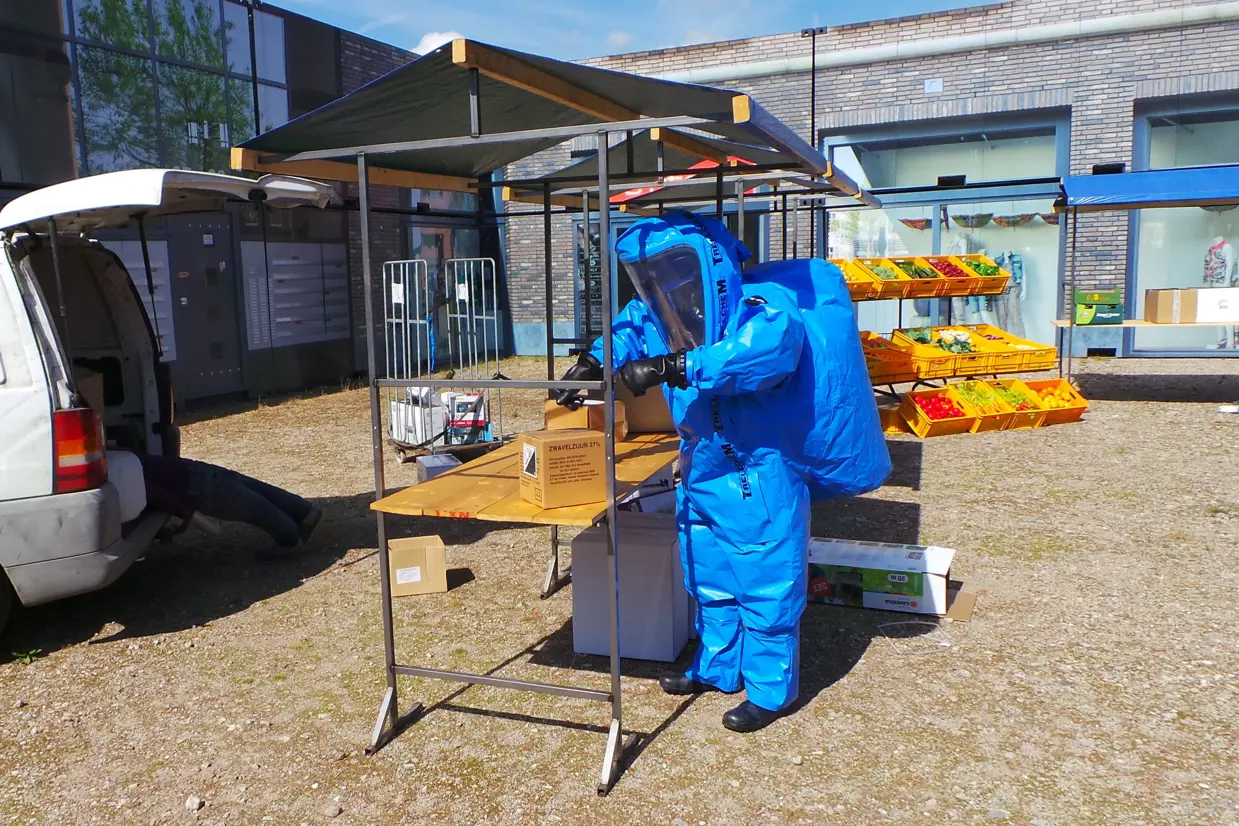 Persoon in beschermend blauw pak onderzoekt een pakketje in een marktkraam.