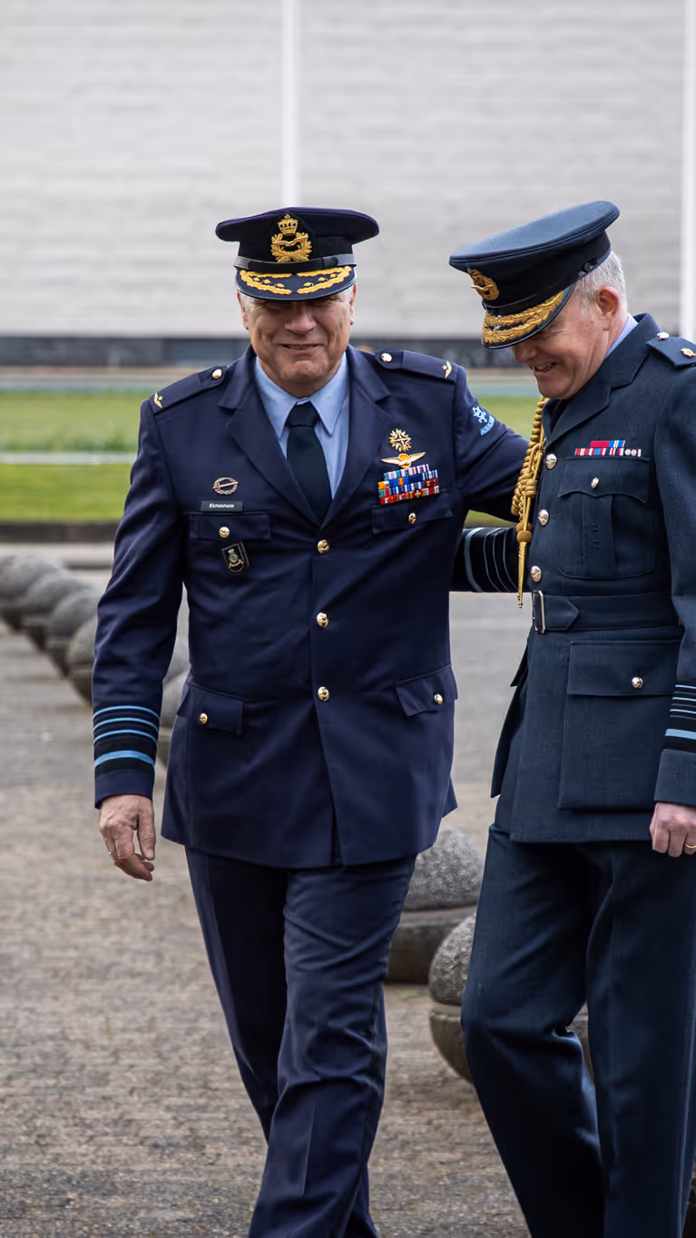 Generaal Onno Eichelsheim loopt lachend naast zijn Britse collega. De twee, in donkerblauw militair uniform, hebben een hand op elkaars rug.