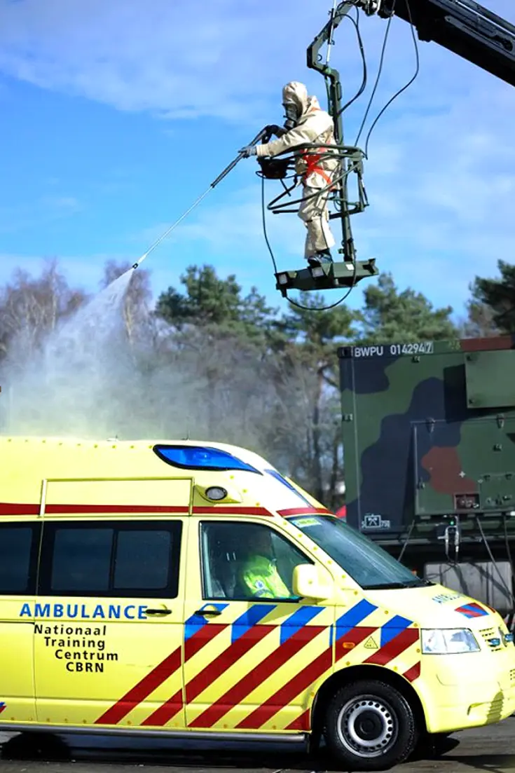 Persoon in beschermend pak spuit vanaf een kraan een ambulance schoon.