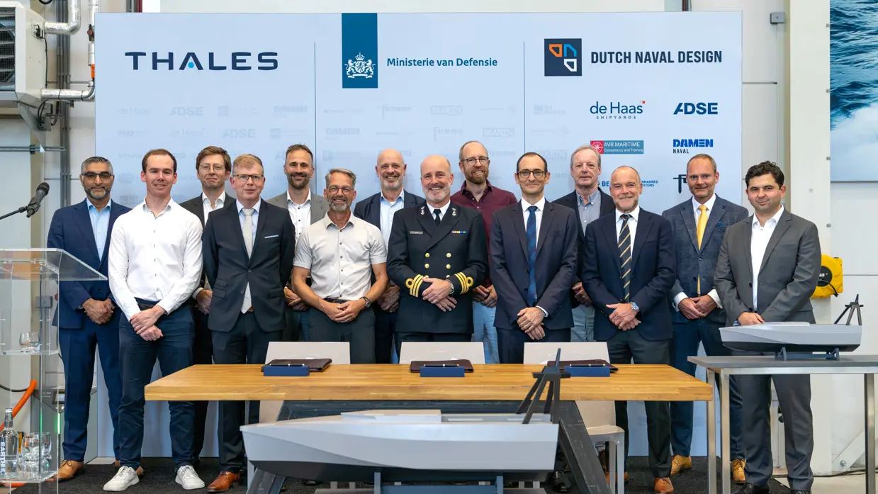 Een grote groep mannen in pak en uniform voor een groot bord met namen van bedrijven.