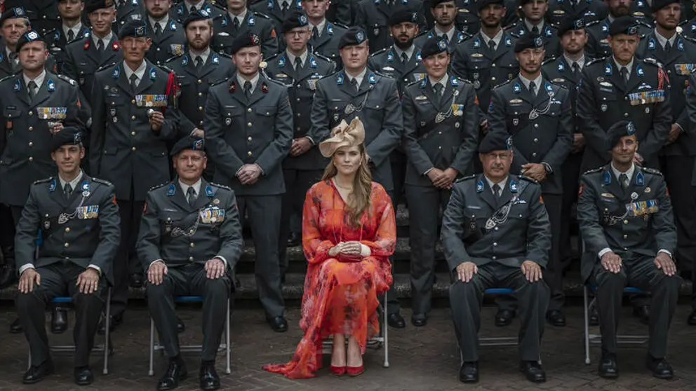 Prinses Amalia in een oranje gewaad zit op een stoel, omringd door landmachtmilitairen in uniform.