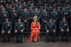 Prinses Amalia in een oranje gewaad zit op een stoel, omringd door landmachtmilitairen in uniform.