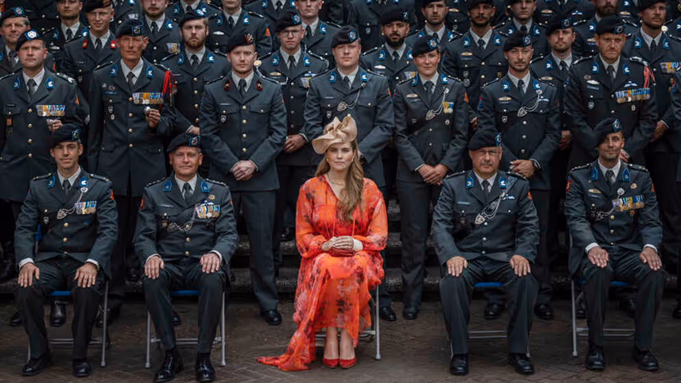 Prinses Amalia in een oranje gewaad zit op een stoel, omringd door landmachtmilitairen in uniform.