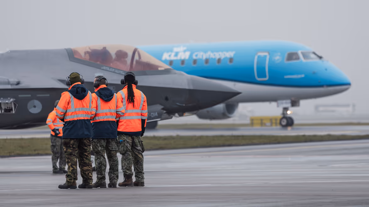 Vier mensen in een oranje veiligheidshesje kijken naar een geparkeerde F-35, waarachter een geparkeerd passagiersvliegtuig van KLM staat. 