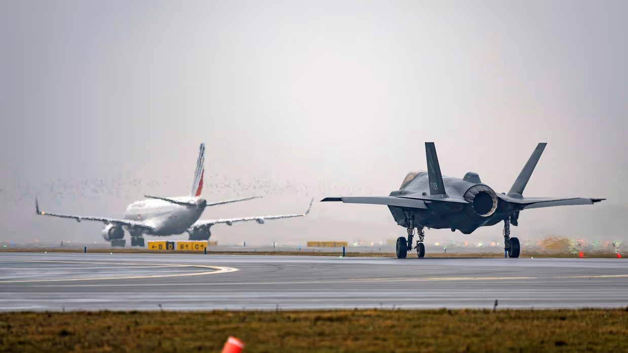 Een taxiënde F-35 rijdt over de landingsbaan op Schiphol. In de verte is een passagiersvliegtuig te zien. De lucht is grijs.