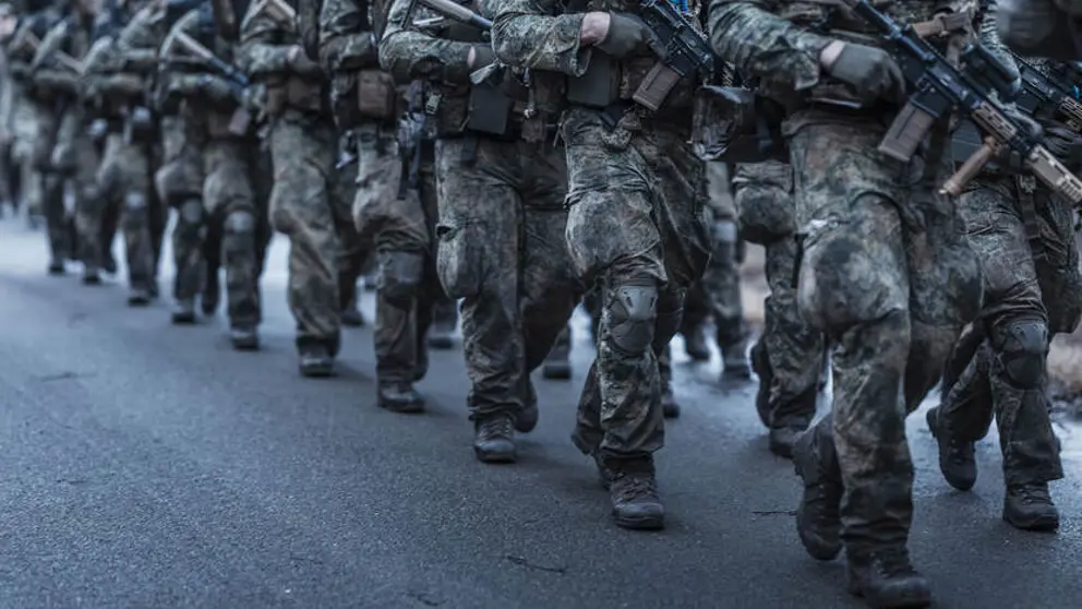 2 rijen marsende deelnemers aan de Nationale Weerbaarheidstraining in uniform met wapen in de hand.