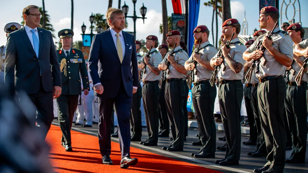 De koning loopt over een oranje loper in de zon en passeert een rij militairen met geweer.