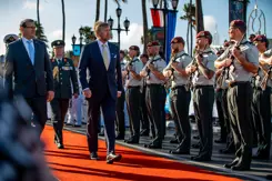 De koning loopt over een oranje loper in de zon en passeert een rij militairen met geweer.