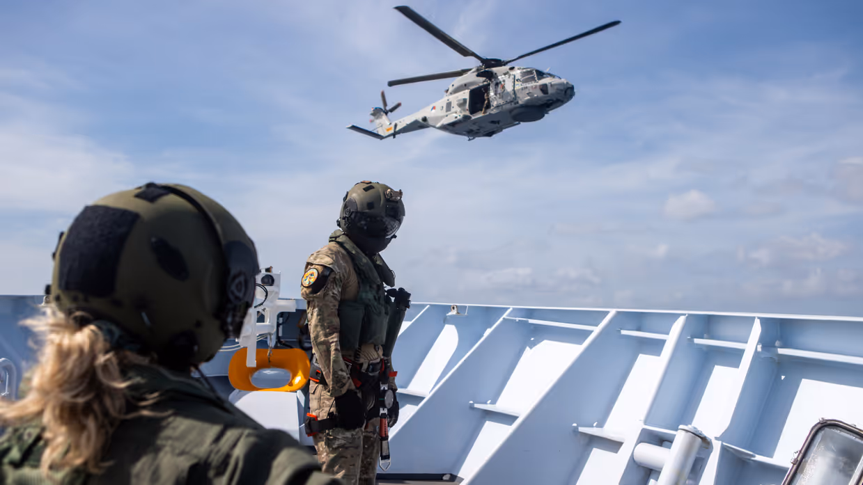 Militairen staan op dek van schip met helikopter in de lucht op de achtergrond.