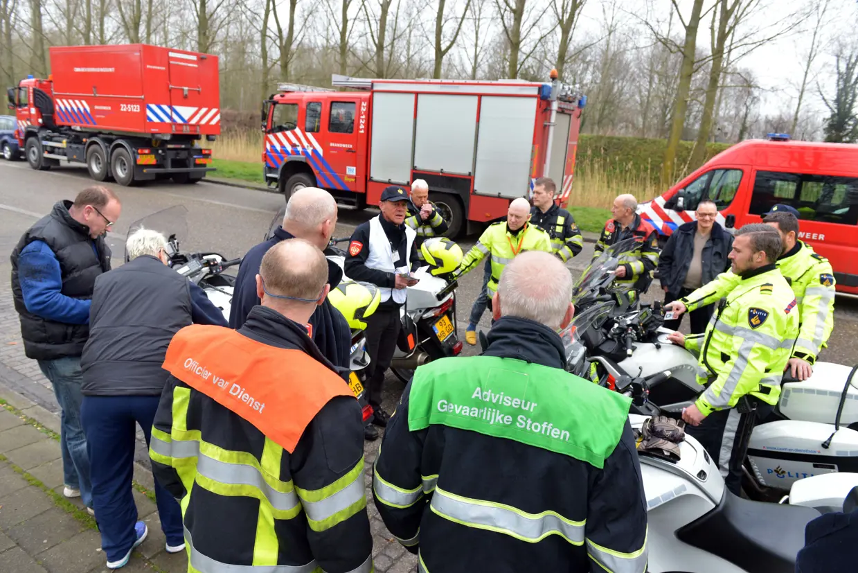 Hulpverleners overleggen in een cirkel. Sommige zitten op een motor. Op de achtergrond staan brandweerauto's.