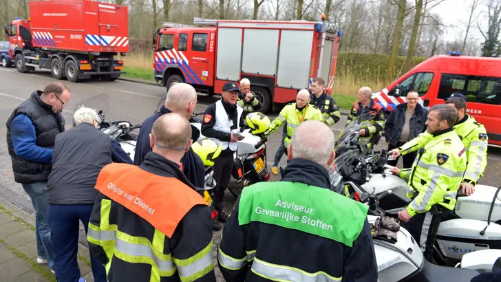 Hulpverleners overleggen in een cirkel. Sommige zitten op een motor. Op de achtergrond staan brandweerauto's.