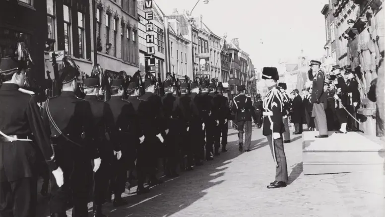 Een zwart-witfoto van een militaire parade met soldaten in uniform, marcherend door een straat met oude gebouwen en toeschouwers.