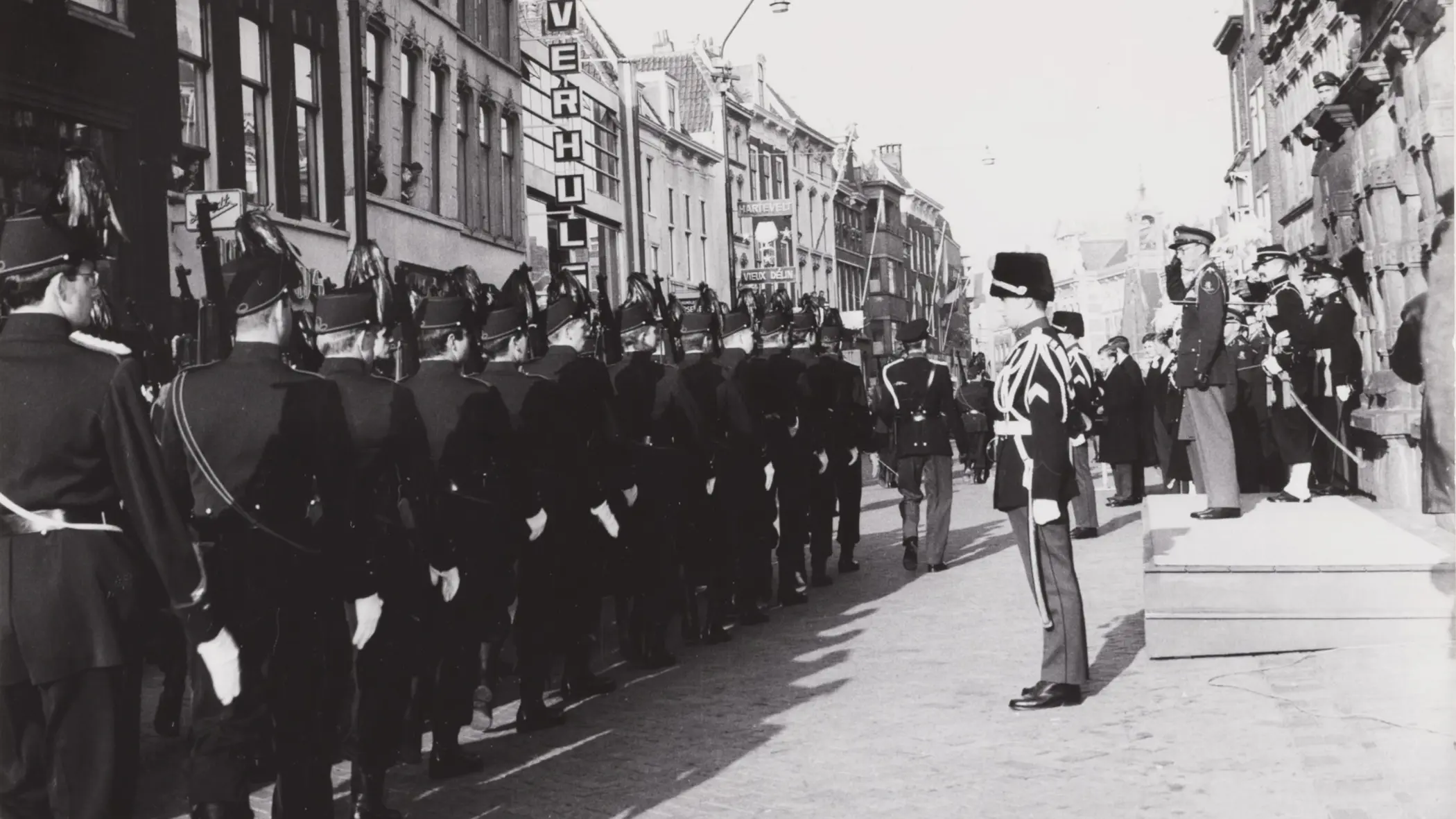 Een zwart-witfoto van een militaire parade met soldaten in uniform, marcherend door een straat met oude gebouwen en toeschouwers.