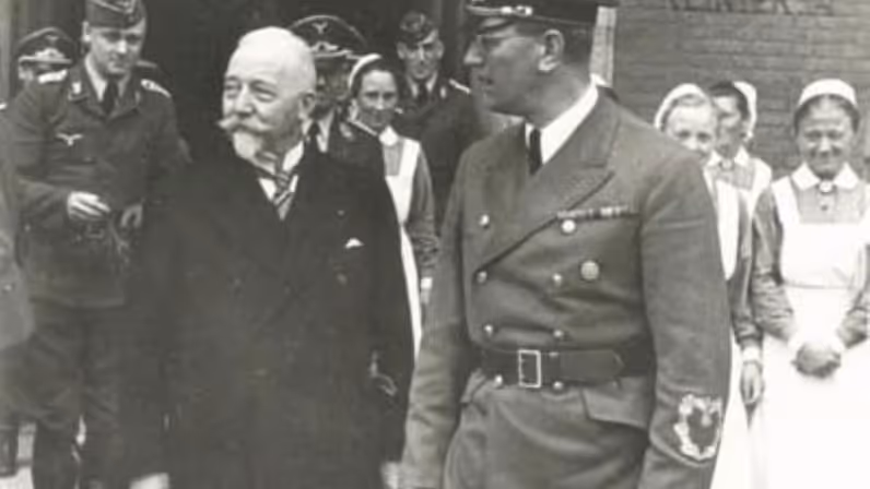 Een oudere man met snor en een man in militair uniform lopen samen, omringd door soldaten en verpleegsters bij een gebouw.