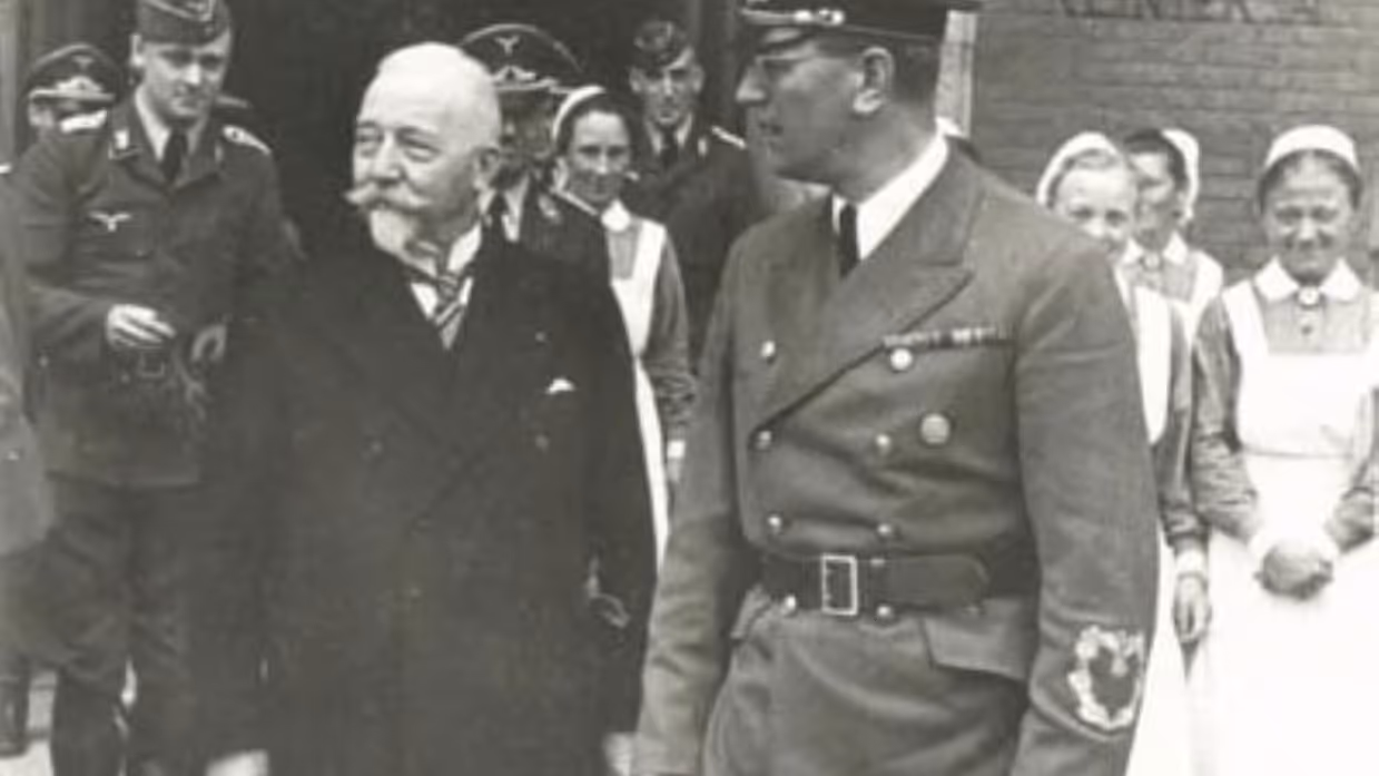 Een oudere man met snor en een man in militair uniform lopen samen, omringd door soldaten en verpleegsters bij een gebouw.