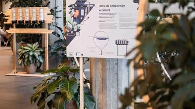  links: 2 informatieborden tussen plantenbakken in een hal. Rechts: Een houten onderzetter met stalen tafelpoot.