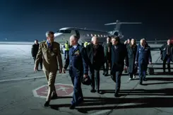 De CDS en minister Brekelmans lopen met een delegatie op een donkere landingsbaan. Op de achtergrond is een vliegtuig te zien.