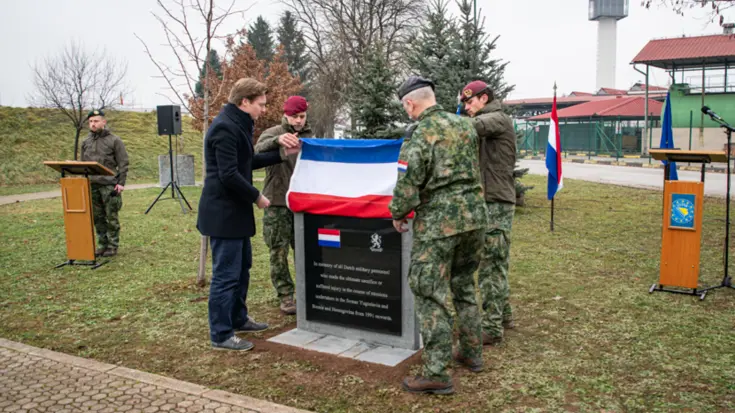 De minister en de CDS halen een Nederlandse vlag omhoog. Daaronder wordt een zwart marmeren gedenkplaat zichtbaar. Ze staan op een grasveld. De lucht is grijs.