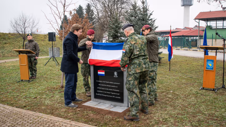 De minister en de CDS halen een Nederlandse vlag omhoog. Daaronder wordt een zwart marmeren gedenkplaat zichtbaar. Ze staan op een grasveld. De lucht is grijs.