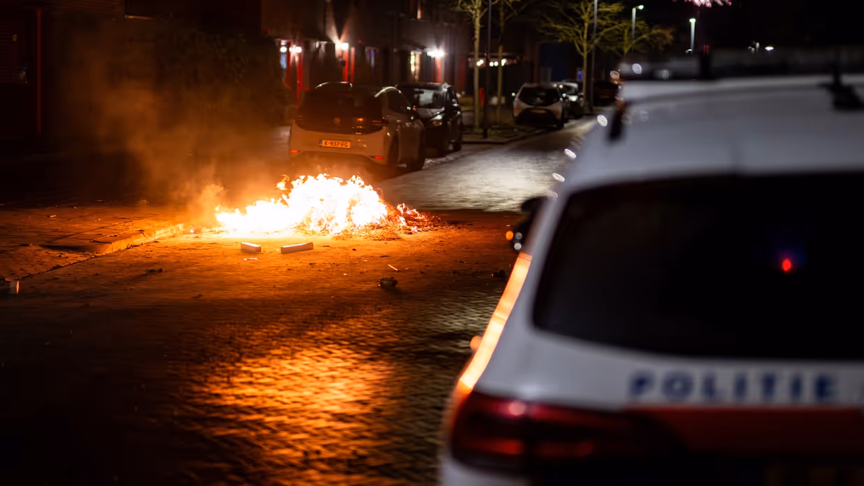 Voertuig van de politie met vlammend vuurwerk op de achtergrond.