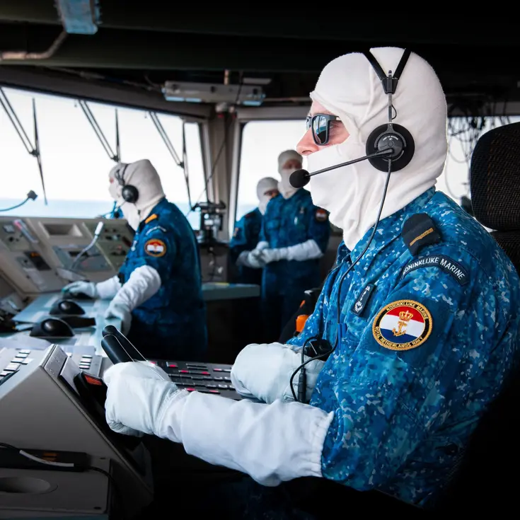 Een marineman zit op de brug van de De Ruyter en draagt antiflash-handschoenen en een antiflash-kap over zijn hoofd.