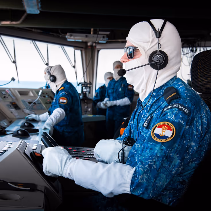 Een marineman zit op de brug van de De Ruyter en draagt antiflash-handschoenen en een antiflash-kap over zijn hoofd.