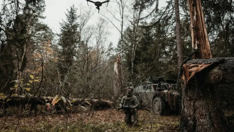 Een militair staat in het bos. Boven hem vliegt een drone die hij bedient.  