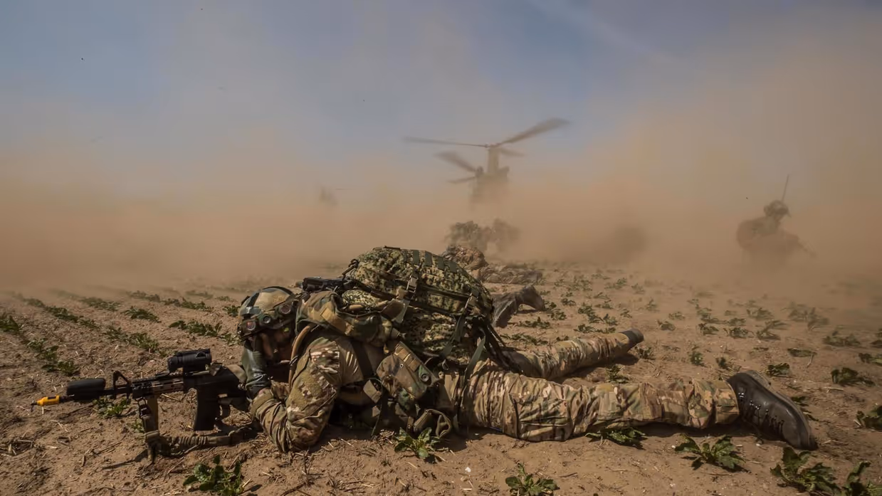 Soldaat ligt in veld met geweer voor zich. Op de achtergrond staat een helikopter met draaiende motor. Zand en stof waait alle kanten op.