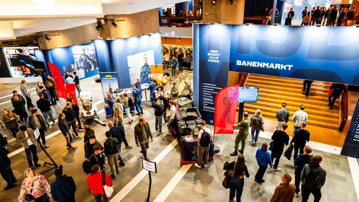 Een volle zaal in bioscoopopstelling met op het podium militairen van de verschillende krijgsmachtdelen en onder meer een groene tent en marechaussee-motor. De zaal is verlicht met roze, oranje en groen.