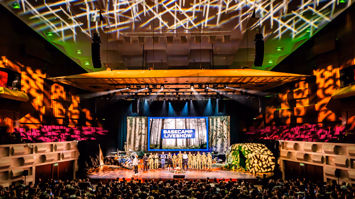 Een volle zaal in bioscoopopstelling met op het podium militairen van de verschillende krijgsmachtdelen en onder meer een groene tent en marechaussee-motor. De zaal is verlicht met roze, oranje en groen.