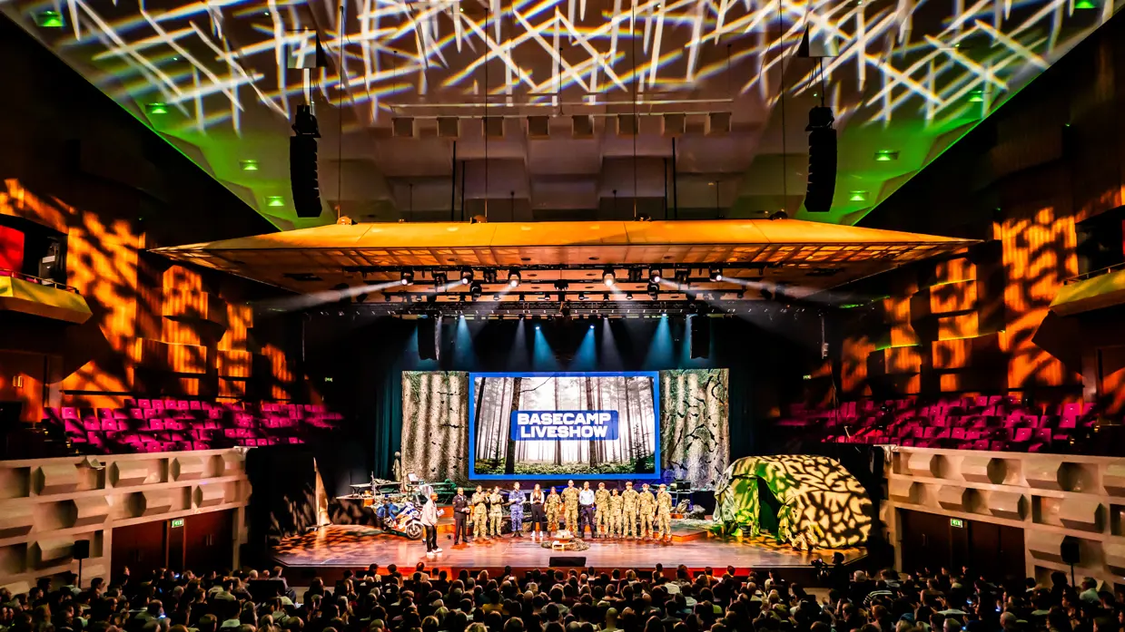 Een volle zaal in bioscoopopstelling met op het podium militairen van de verschillende krijgsmachtdelen en onder meer een groene tent en marechaussee-motor. De zaal is verlicht met roze, oranje en groen.