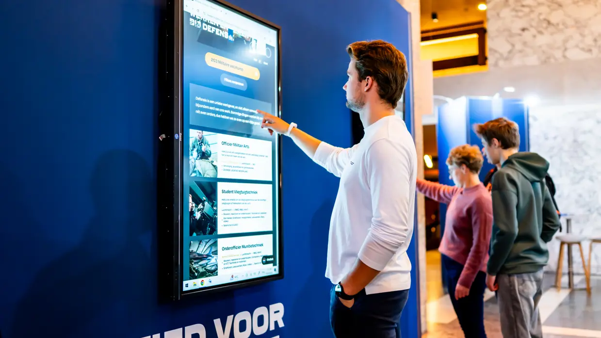 Een volle zaal in bioscoopopstelling met op het podium militairen van de verschillende krijgsmachtdelen en onder meer een groene tent en marechaussee-motor. De zaal is verlicht met roze, oranje en groen.