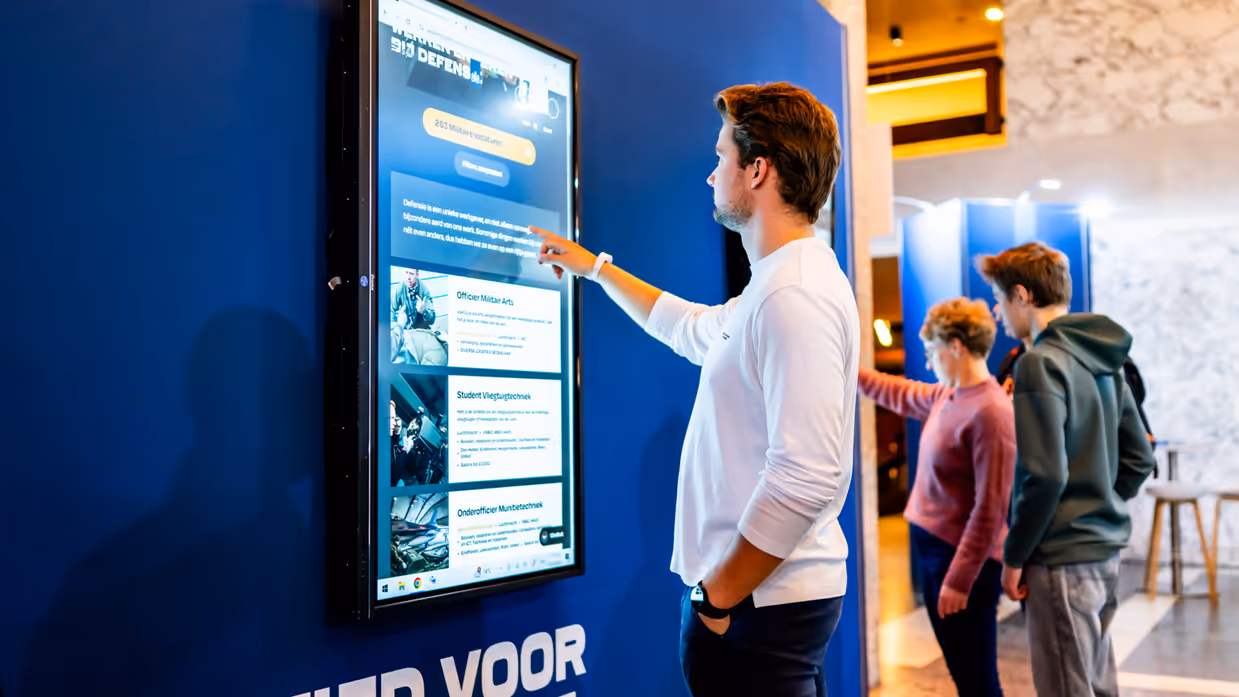 Een volle zaal in bioscoopopstelling met op het podium militairen van de verschillende krijgsmachtdelen en onder meer een groene tent en marechaussee-motor. De zaal is verlicht met roze, oranje en groen.