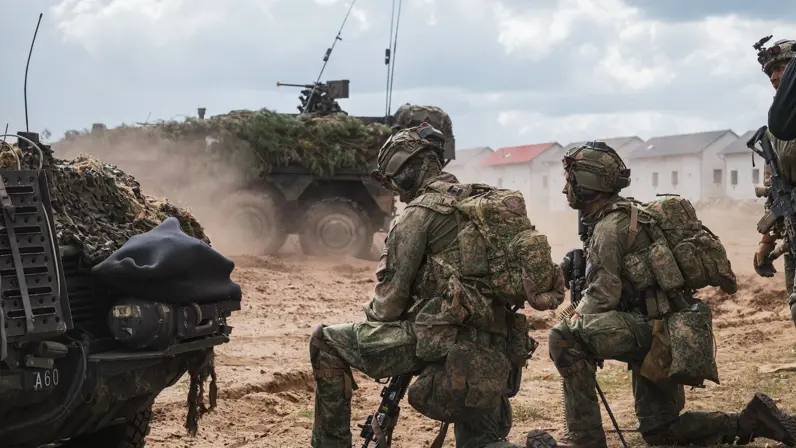 2 omgehangen militairen zitten op een knie, voor hen staat een voertuig en ook achter hen rijdt er een voorbij.&nbsp;