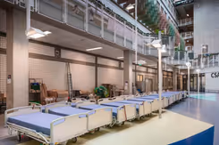 Een dertiental bedden met blauwe matrassen staan in de hal van een ziekenhuis.