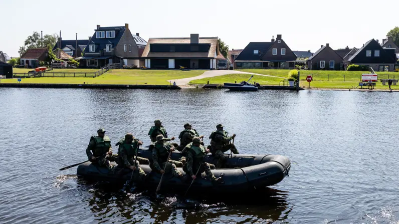 Een zwarte opblaasboot met negen militairen in camouflagekleding vaart over rustig water, met huizen en groen op de achtergrond. 