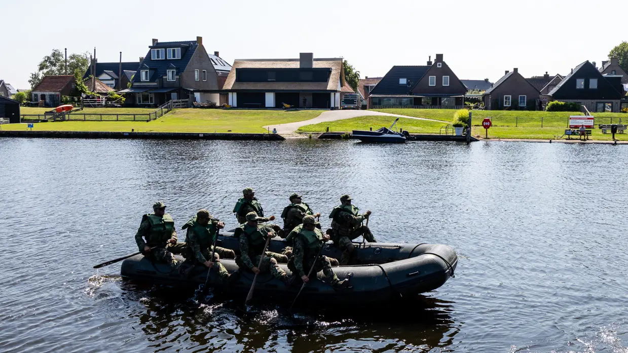Een zwarte opblaasboot met negen militairen in camouflagekleding vaart over rustig water, met huizen en groen op de achtergrond. 