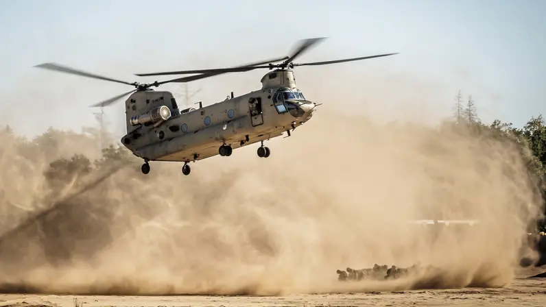 Een militaire helikopter vliegt laag boven een stoffige grond, met zand en stof die opwaaien door de rotorbladen.