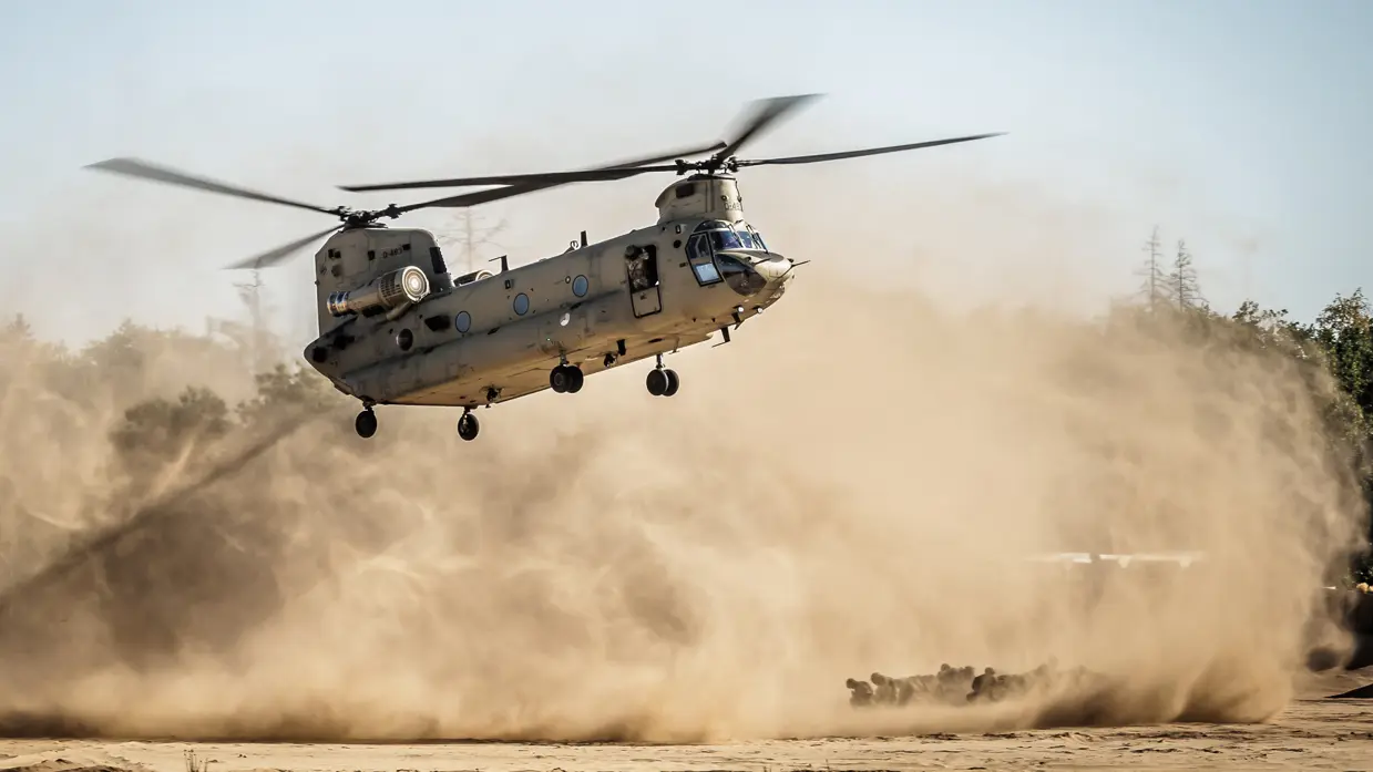 Een militaire helikopter vliegt laag boven een stoffige grond, met zand en stof die opwaaien door de rotorbladen.