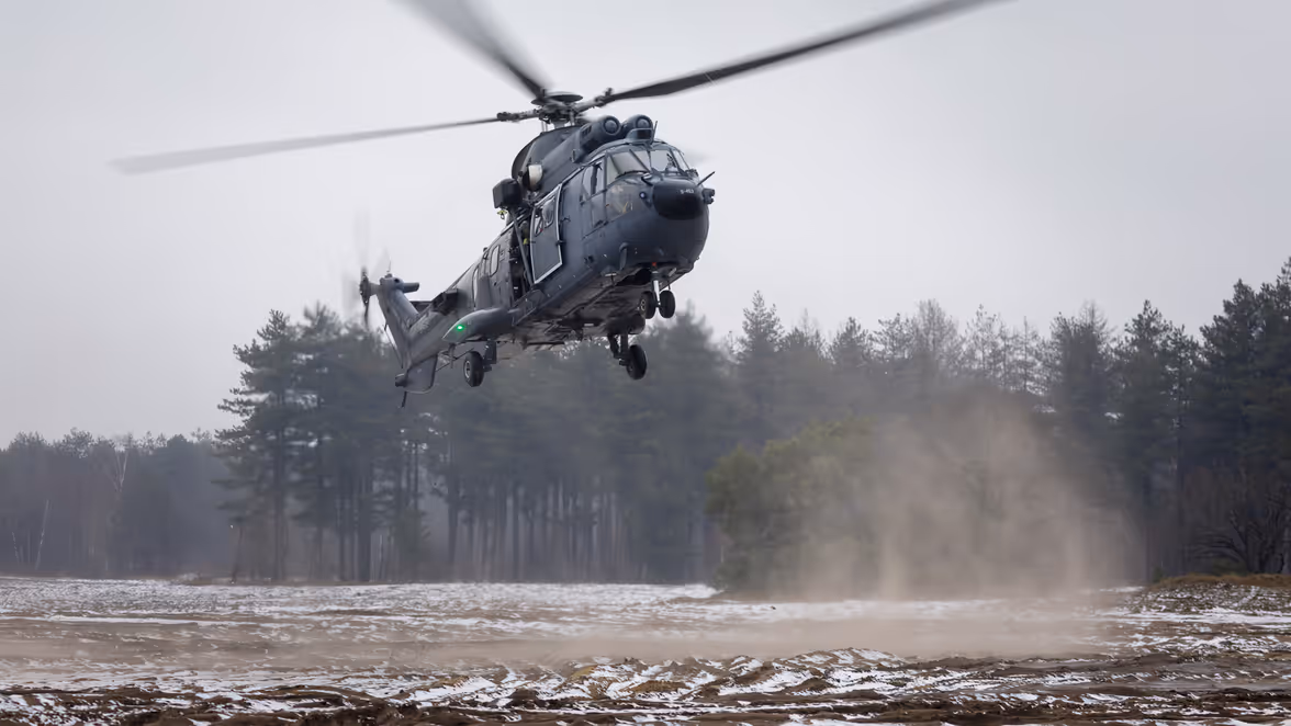  Een militaire helikopter landt boven een veld waarop nog wat sneeuw ligt bij een blauwe hemel.