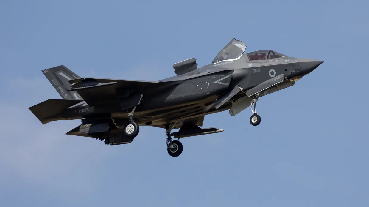  Een zwarte straaljager met open cockpit en ingetrokken wielen, vliegend tegen een heldere blauwe lucht met zichtbare markeringen en nummers.    